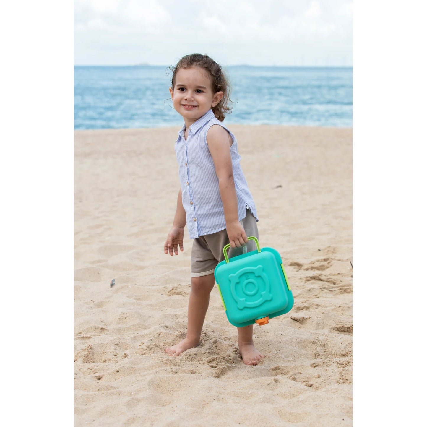 Child holding a teal sandcastle mold on a sandy beach with ocean in the background