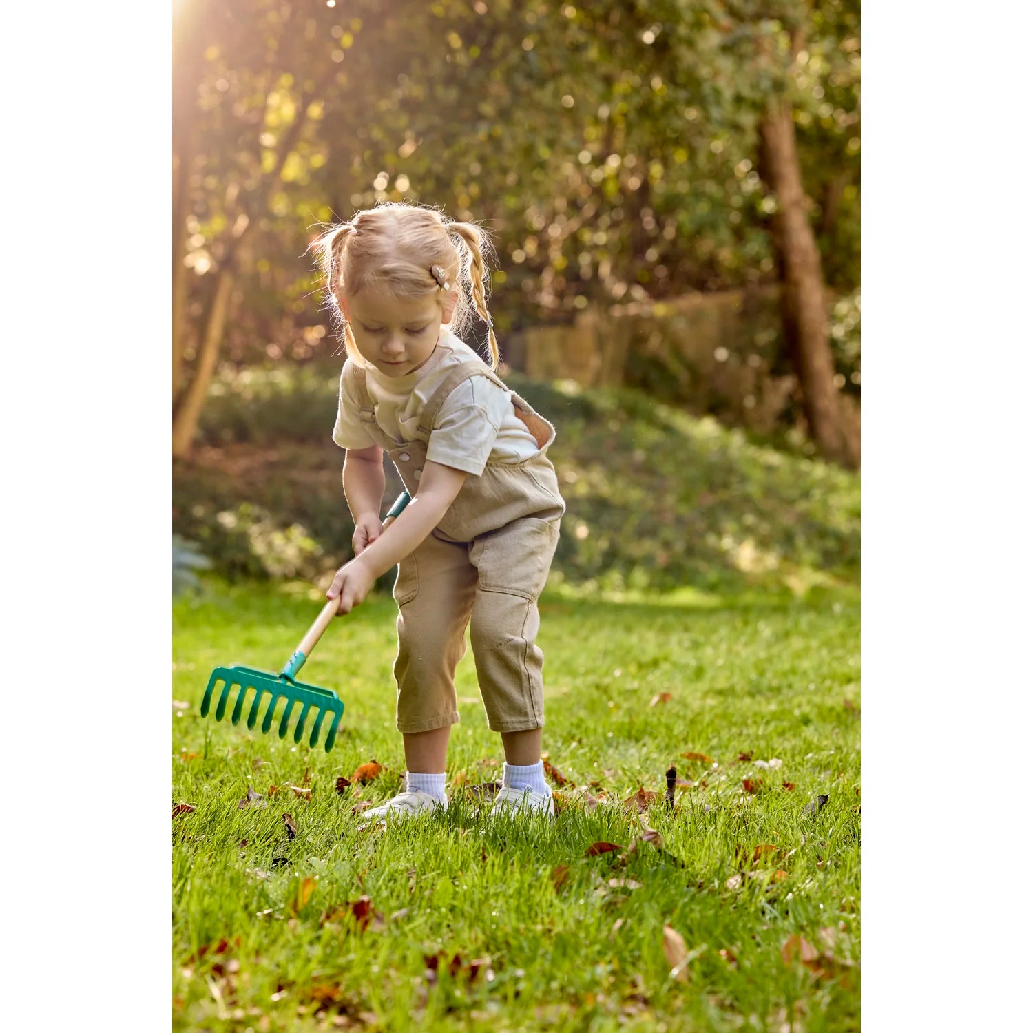 Child in a garden using a green rake on a grassy area with trees in the background.