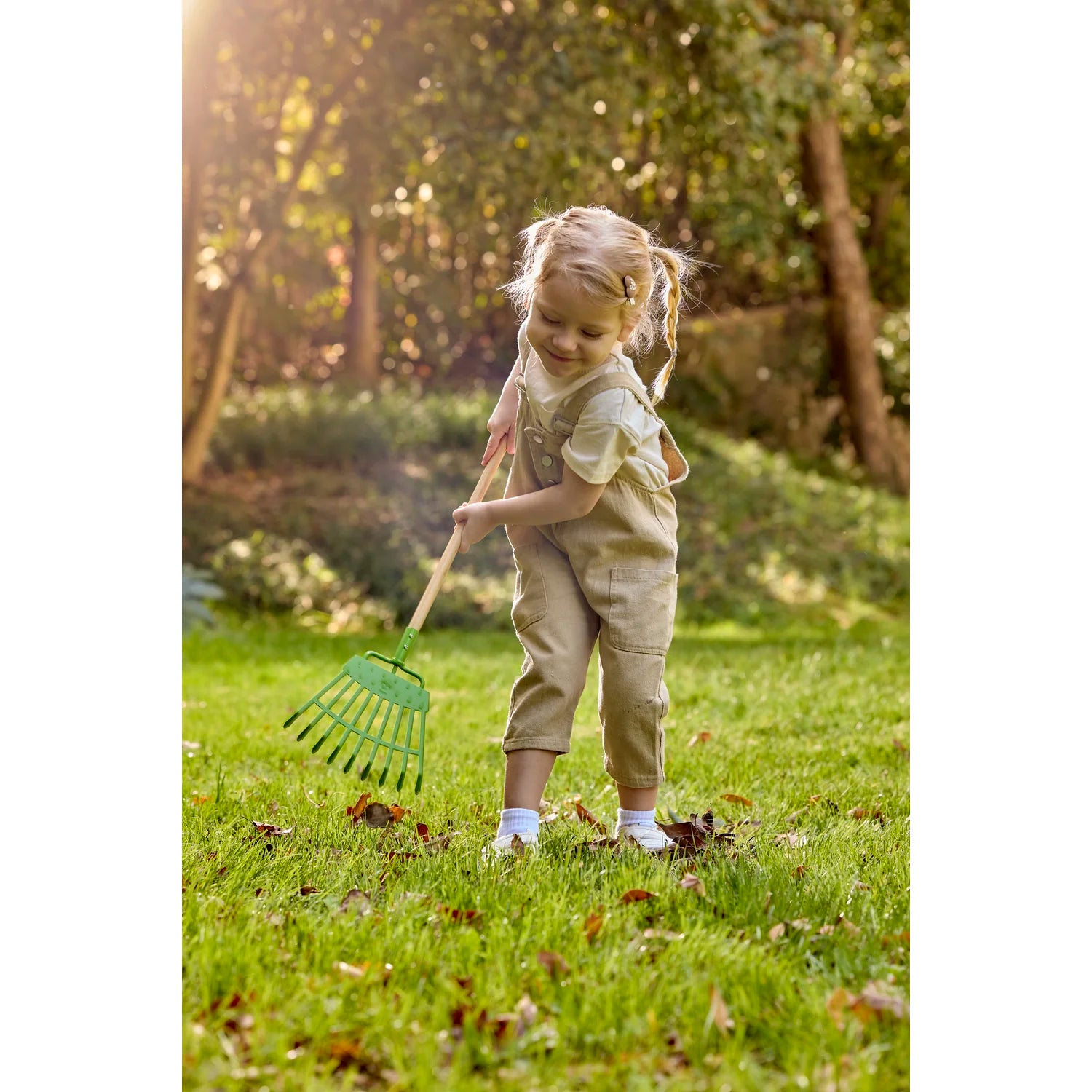 Child playing with a green garden rake in a grassy area with trees in the background