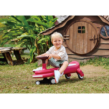 Child playing on a pink toy car in a garden with a wooden playhouse in the background