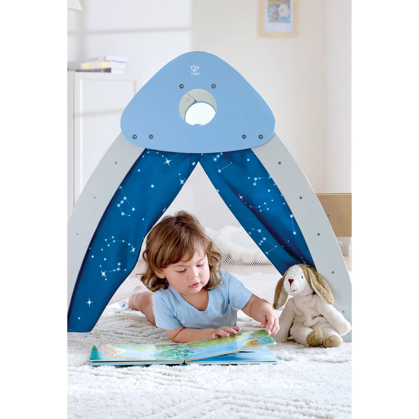 Child reading a book under a blue and white tent with star patterns, surrounded by toys.
