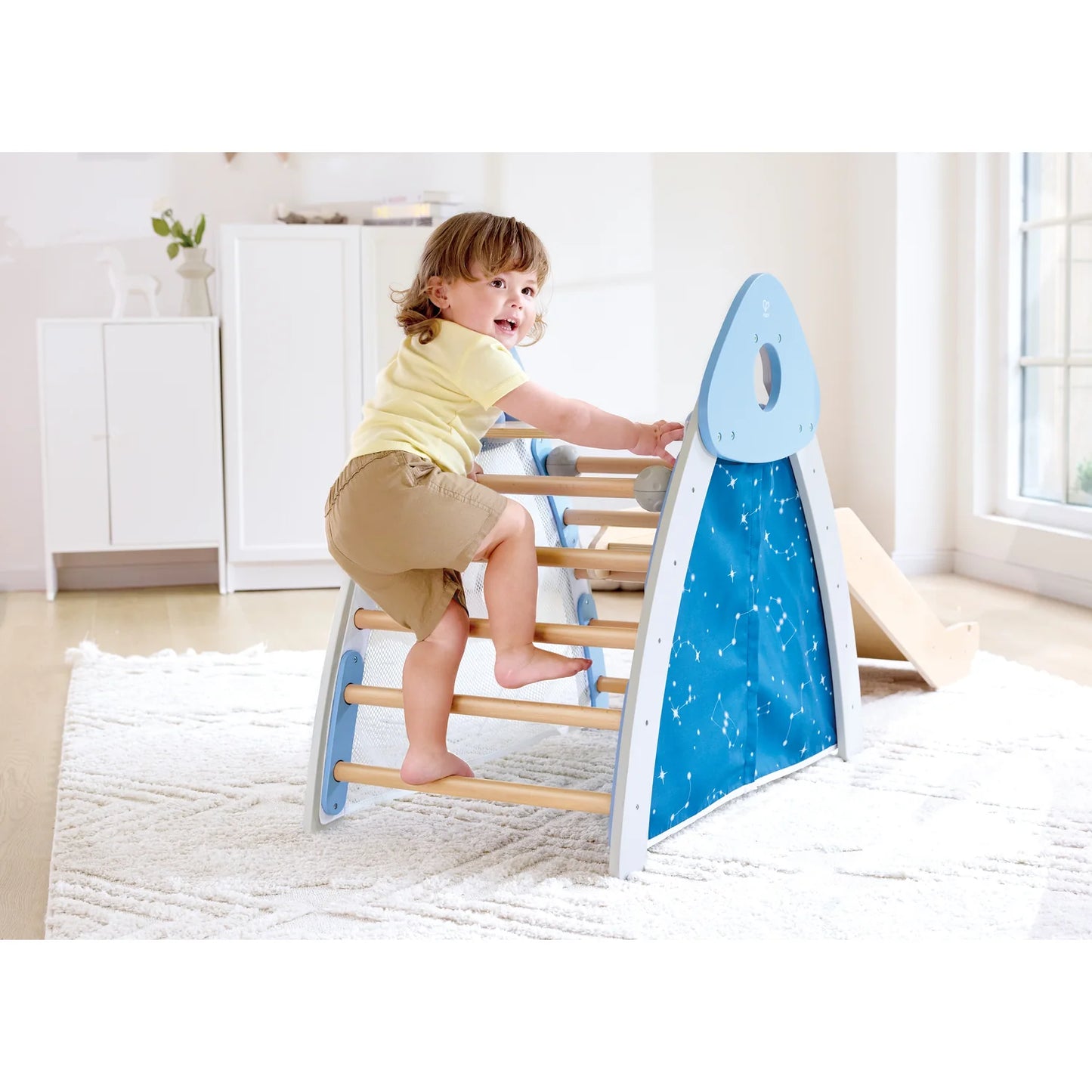 Child climbing on a blue and wooden slide in a room with white walls and a window.