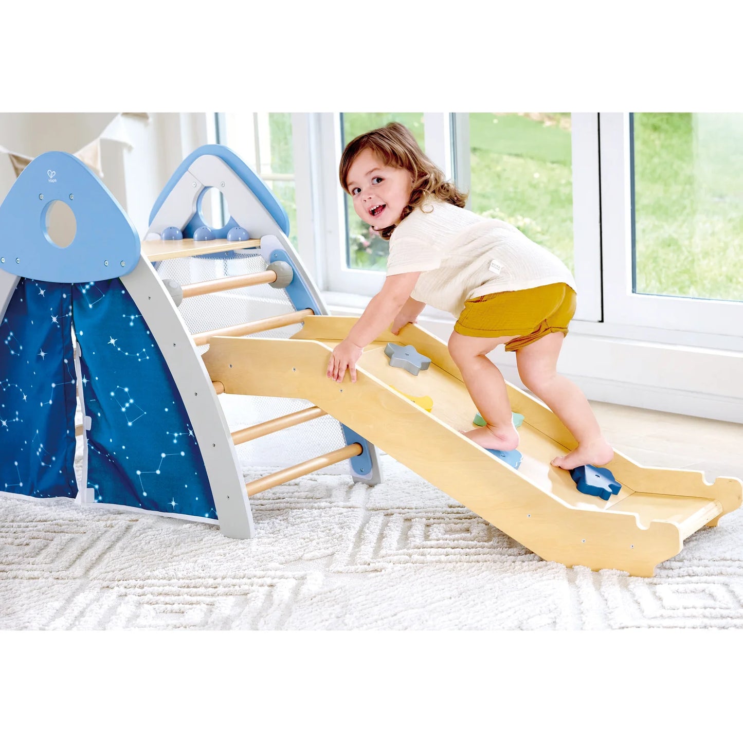 Child playing on a yellow slide in a play area with a blue curtain and white walls.