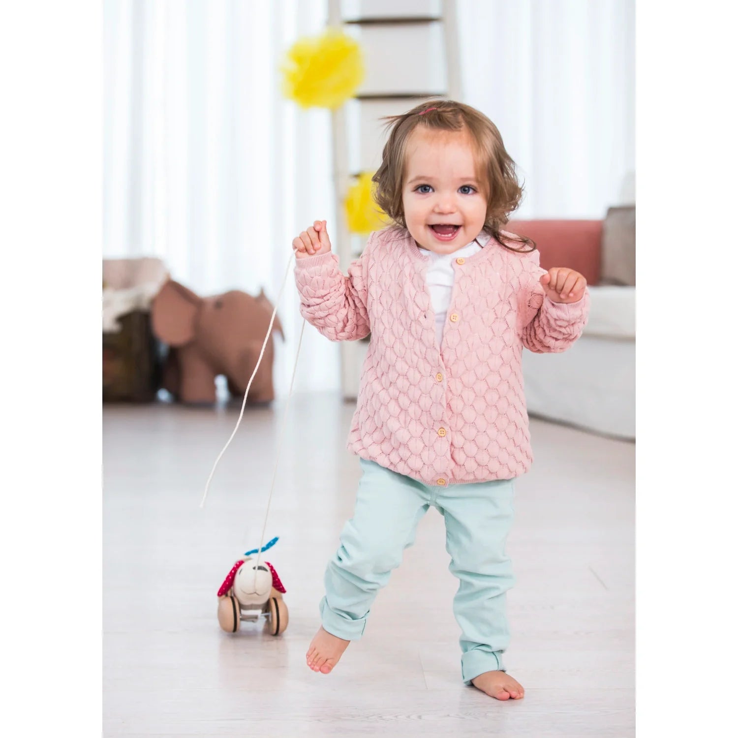 A toddler walking with a pink push and pull along dog toy with wheels. The toy is made of wood with a soft fabric finish, and it's designed for children to pull along the dog on a leash.