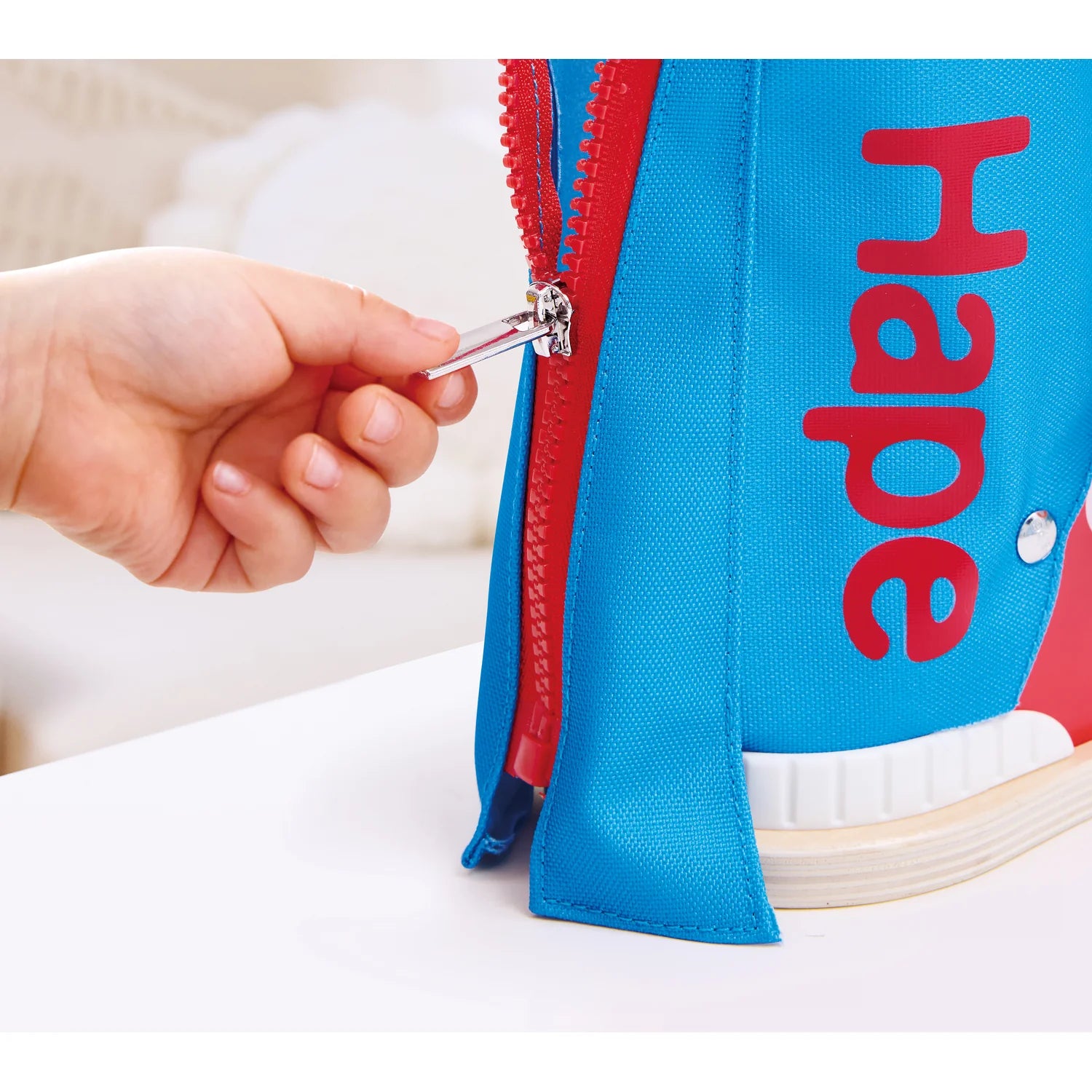 Hand zipping a blue bag with red zipper and 'hape' branding.