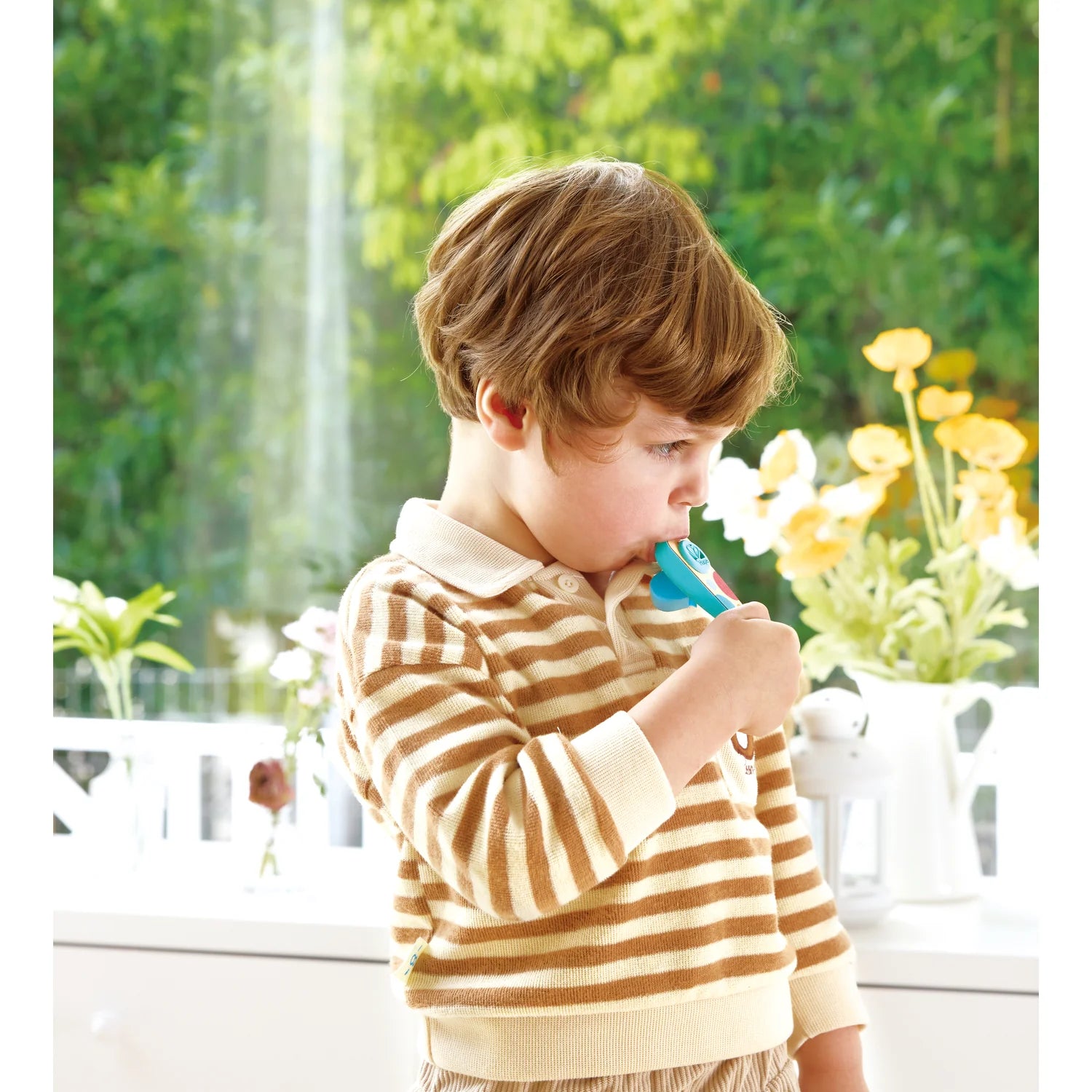 Child holding a blue toy in front of a window with greenery outside