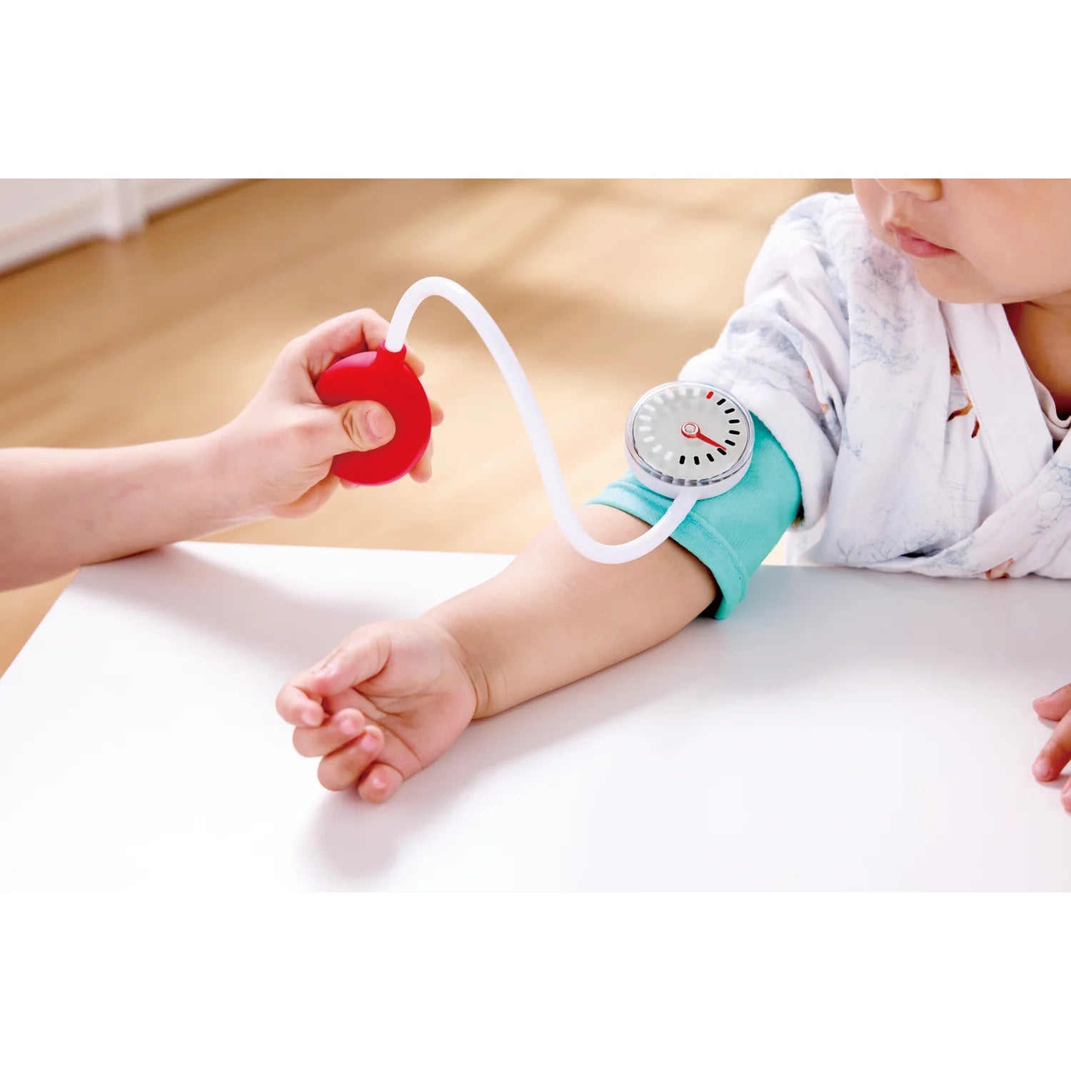 Baby's arm with a blood pressure monitor and a hand holding a red device on a light-colored surface.