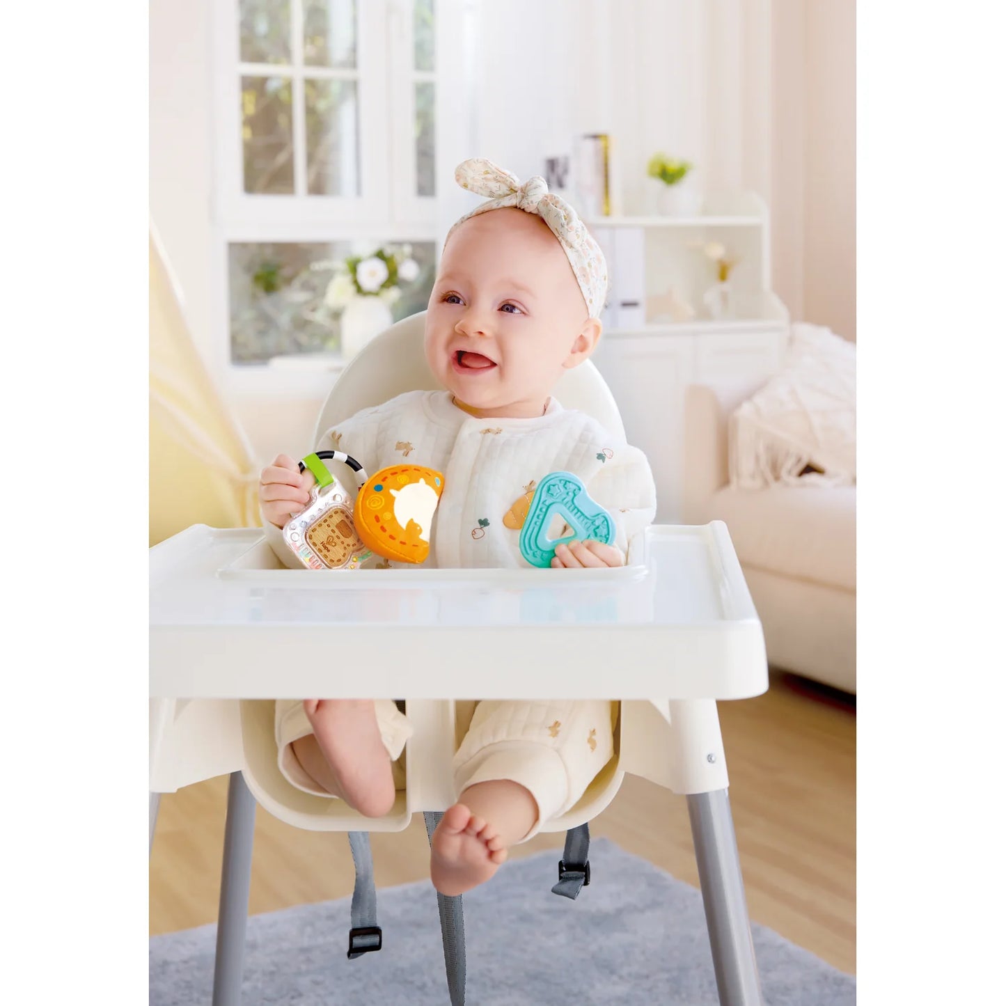 Baby in a high chair with toys, sitting in a bright room.