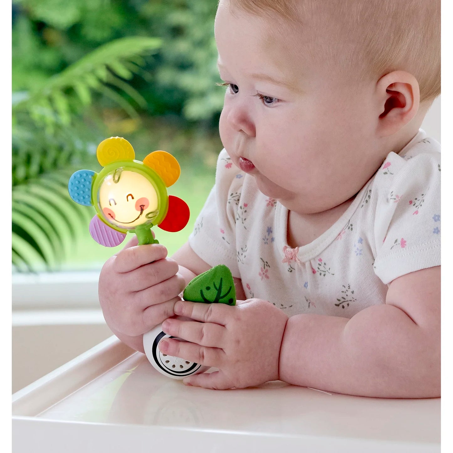 Baby holding a colorful toy with a smiling face, sitting on a white surface with greenery in the background.