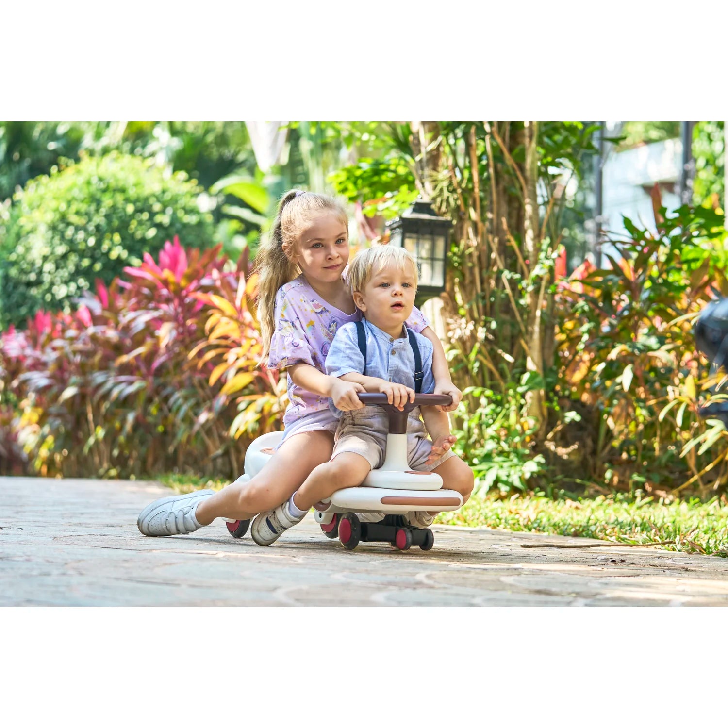 Two children riding a scooter in a garden setting with greenery and flowers.