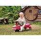 Child playing on a pink toy car in a garden with a wooden playhouse in the background