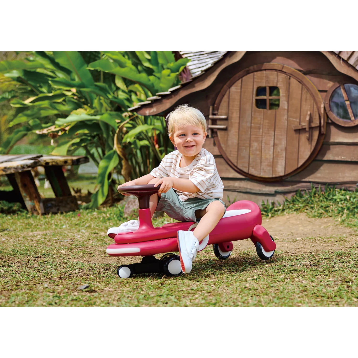 Child playing on a pink toy car in a garden with a wooden playhouse in the background